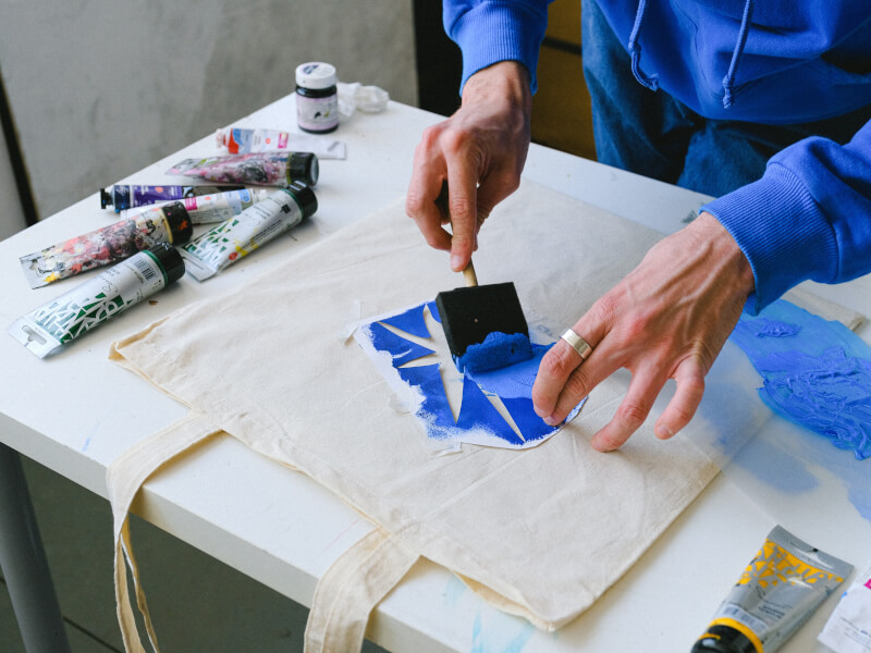 A person is printing a design on a tote bag in blue ink using a stencil and sponge